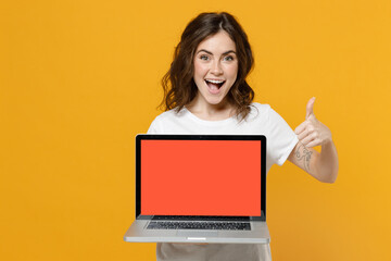 Naklejka premium Young fun freelancer copywriter student woman in white basic t-shirt hold laptop pc computer with blank screen workspace area show thumb up like gesture isolated on yellow background studio portrait.