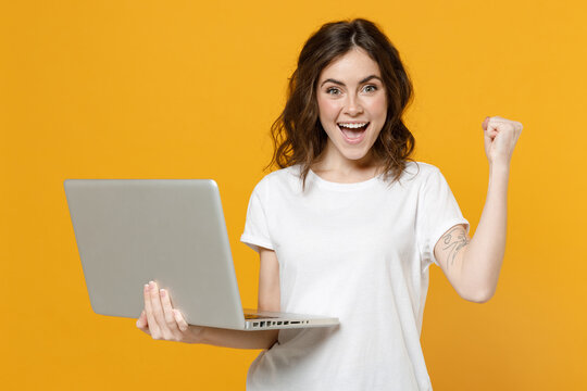 Young smiling freelancer copywriter student woman in white basic t-shirt hold laptop pc computer browsing do winner gesure clench fist celebrating isolated on yellow orange background studio portrait.