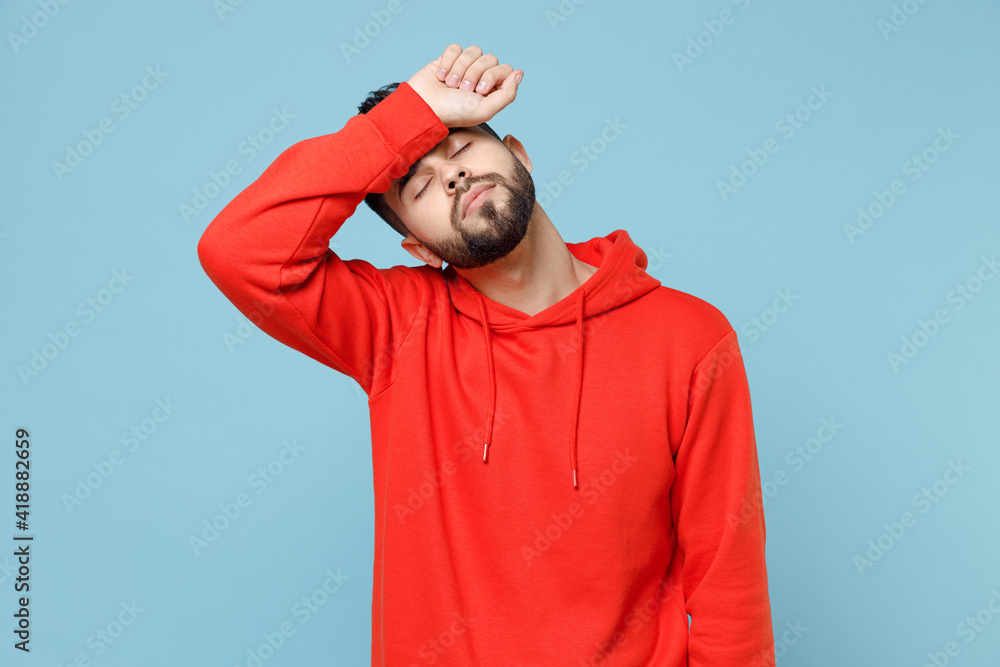 Wall mural Young caucasian tired exhausted sick ill student bearded man 20s in casual red orange hoodie put hand on forehead having headache isolated on blue background studio portrait People lifestyle concept. - Wall murals