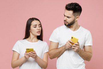 Young shocked couple two friends man woman in white basic blank print design t-shirts hold in hand using mobile cell phone pry into chatting typing sms spy on peep isolated on pastel pink background.