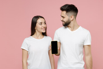 Young cheerful couple two friends man woman 20s in white basic blank print design t-shirt hold in hand mobile cell phone with with blank black screen workspace area isolated on pastel pink background