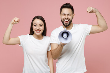 Young fun couple two friends man woman in white basic blank print design t-shirts hold scream in megaphone announces discounts sale Hurry up isolated on pastel pink color background studio portrait.