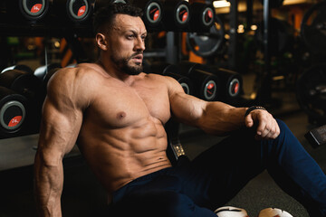 Fototapeta na wymiar A male athlete with a beautiful naked body sits on the gym floor near the dumbbell and looks straight. Photos for advertising fitness center. High quality photo