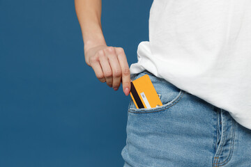 Close up cropped photo portrait shot of caucasian female hand arm putting credit bank card in jeans pants denim pocket isolated on dark blue background studio portrait. Money finance currency concept. - obrazy, fototapety, plakaty