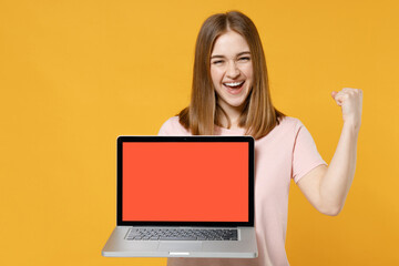 Naklejka premium Young freelancer woman 20s in casual basic pastel pink t-shirt holding laptop pc computer with blank screen workspace area do winner gesture clench fist isolated on yellow background studio portrait..