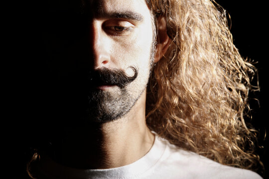 Uomo con baffi curati e capelli lunghi guarda verso il basso con espressione affranta, isolato su sfondo scuro