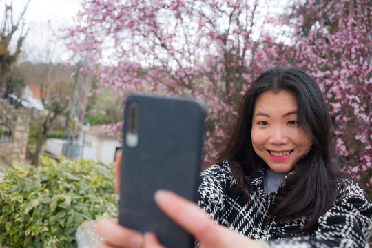 spring sakura selfie - young happy and cute Asian Korean tourist woman taking self portrait with mobile phone smiling cheerful in front of blooming trees  in beautiful pink colors