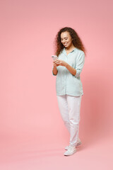 Obraz premium Full length young black african fun happy smiling curly student woman 20s in blue shirt holding mobile cell phone chatting browsing internet isolated on pastel pink color background studio portrait.
