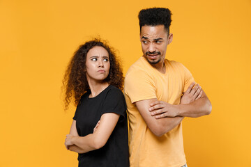 Young couple friends together team african resentful frowning man woman in black tshirt stand back to back with hands crossed folded look to each other isolated on yellow background studio portrait