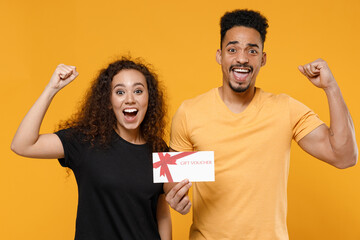 Young couple two friends together family african overjoyed smiling happy man woman in black t-shirt hold gift voucher flyer mock up do winner gesture clench fist isolated on yellow background studio.