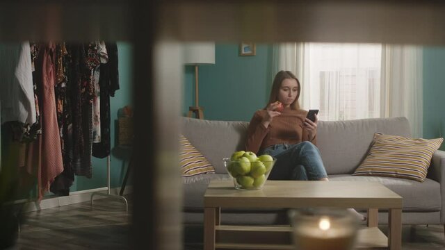 Young woman in blue jeans and brown sweater sits on beige sofa on window background, bit red apple and choked. Trying to cough. On table is glass jar with green apples. Camera dollies horizontally