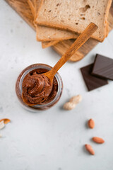 Fototapeta na wymiar Chocolate nut paste spread in a jar and on a wooden spoon with peanuts and breakfast bread