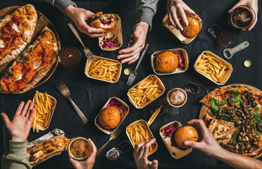 Lockdown fast food dinner from delivery service. Flat-lay of peoples hands eating burger, fries, sandwiche, pizza, drinking beer over table background, top view. Quarantine home party, takeaway food