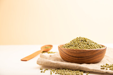 Wooden bowl with raw green mung bean and wooden spoon on a white and orange background. Side view, selective focus, copy space.