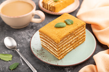 Homemade honey cake with milk cream and mint with cup of coffee on a black concrete background. Side view, selective focus.