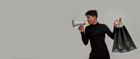 Stylish young guy dressed elegantly in black screaming in megaphone, making sale announcement, posing with shopping bags isolated over gray background