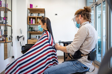 Young woman have hair cutting at hair stylist during pandemic isolation, they both wear protective equipment. Hair dresser working with a face mask during coronavirus quarantine