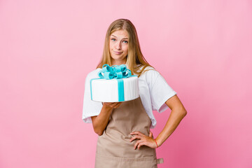 Young russian baker woman holding a delicious cake unhappy looking in camera with sarcastic expression.