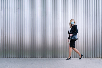 Elegant business woman with face mask moves with her handbag on the street on metallic background. Horizontal copy space