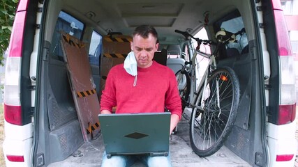 Communication of a male loader in the trunk of a car. He speaks into the phone. Uses a laptop to process digital data. Cargo transportation service. - Powered by Adobe
