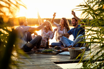 Group of friends having fun on picnic near a lake, sitting on pier eating and drinking wine.