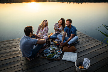 Group of friends having fun on picnic near a lake, sitting on pier eating and drinking wine.