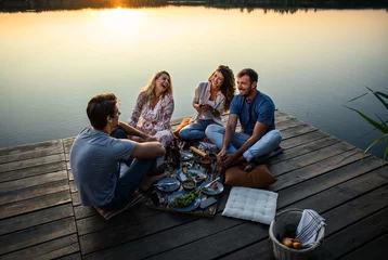 Fotobehang Group of friends having fun on picnic near a lake, sitting on pier eating and drinking wine. © Zoran Zeremski