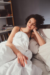 Woman lying in white bed on pillows under blanket, resting head on hands