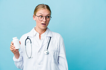 Young russian doctor woman holding pills bottle on blue being shocked because of something she has seen.