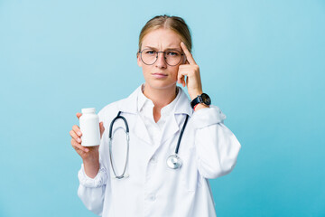 Young russian doctor woman holding pills bottle on blue focused on a task, keeping forefingers pointing head.