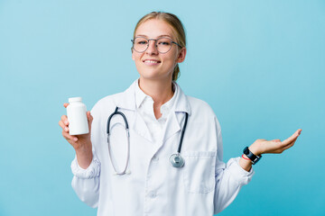 Young russian doctor woman holding pills bottle on blue showing a welcome expression.