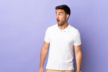 Young handsome man isolated on purple background doing surprise gesture while looking to the side