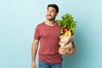 Young caucasian man buying some vegetables isolated on blue background laughing and looking up