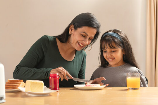 AN ADULT WOMAN SPREADING JAM ON BREAD FOR HER DAUGHTER SITTING BESIDE	