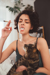 Woman is holding striped cat with big green eyes while smoking marijuana at home