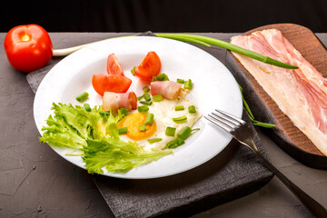 Scrambled eggs with bacon, garnished with tomato and green onion and sliced bacon and tomato and onion on a gray concrete background on a wooden stand.