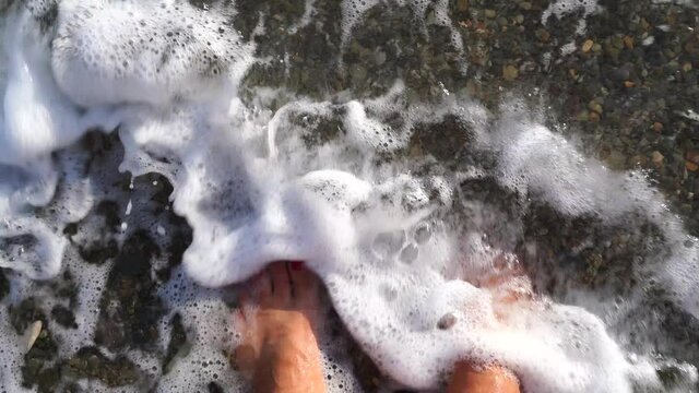 A sea wave washes over my bare feet. Summer freshness of the sea wave. A wave covered the woman's legs with a red pedicure. The sea wave washes the sand from your feet.