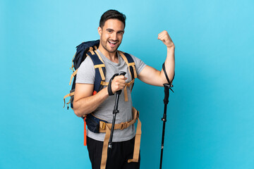 Young caucasian man with backpack and trekking poles isolated on blue background doing strong gesture