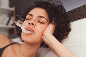 Enjoyment. Close-up of woman smoking weed and blowing puffs of smoke while laying at the bed
