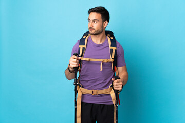 Young caucasian man with backpack and trekking poles isolated on blue background looking to the side