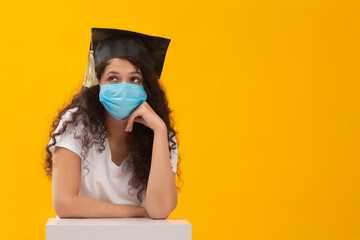 A TEENAGER WEARING FACE MASK AND GRADUATION CAP SITTING AND THINKING	