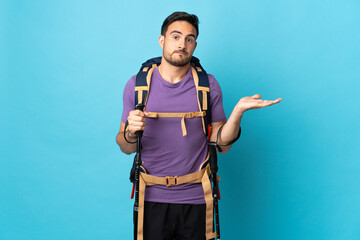 Young caucasian man with backpack and trekking poles isolated on blue background having doubts