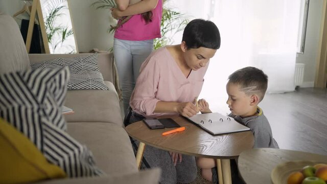 careful brunette mother playing or do lessons with little preschool age boy in paper notebook at small table while teenage girl plays with kitten on floor in living room