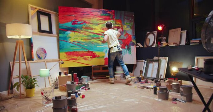 Joyful Caucasian young talented male artist listening to favorite song and dancing while drawing oil painting on big canvas in cozy studio. Creative man painter creating abstract artwork in workshop