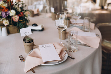 Wedding banquet. The festive table is decorated with compositions of flowers and greenery. On the table are glasses, talers with napkins, cutlery and name cards and menu cards.