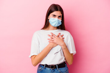 Young caucasian woman wearing a mask for virus isolated on pink background has friendly expression, pressing palm to chest. Love concept.