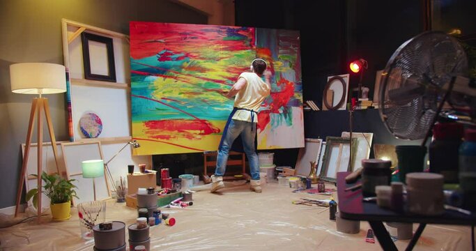 Rear of Caucasian young talented male artist drawing oil painting on big canvas standing in art studio listening to favorite music and dancing. Creating abstract artwork in workshop, painter concept
