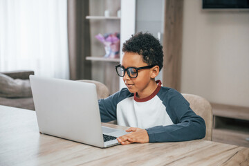 Aftican american child in glasses sits by table with laptop at home