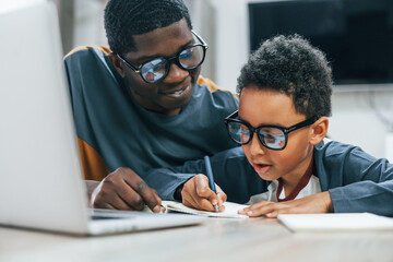 African american father with his son is indoors at home. Using laptop. Conception of education