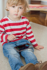 Cute little child using a smartphone while sitting on the floor at home. Digital device and screen time addiction, technology in the hands of children concept.